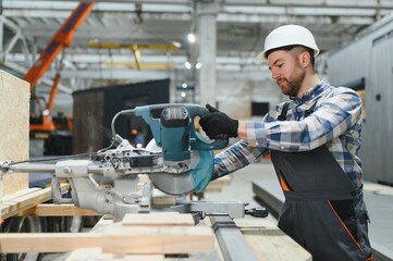 A carpenter cuts a wooden plank on a sawmill in a modular building factory, providing precise cuts...