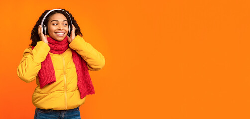 Happy African Lady Listening To Music Enjoying Song And Sound In Wireless Earphones Posing Standing In Studio Over Yellow Background, Wearing Winter Clothes. Autumn Playlist Concept. Copy Space