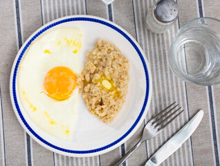 Boiled oatmeal with a piece of butter and a fried egg on a white plate, side view. Serving breakfast on a gray table