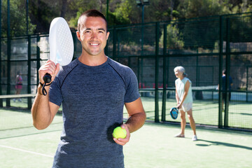 Portrait of a satisfied adult man with racket and ball in his hands on the padel court