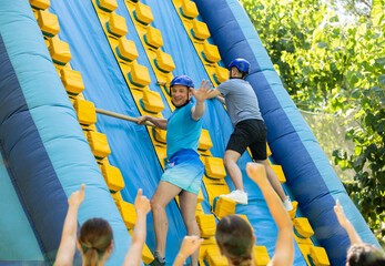 Happy active sporty adult man having funny competition in climbing on inflatable castle with wooden sticks in summer outdoor amusement park..