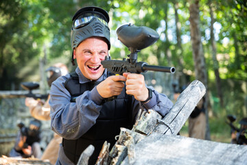 Fototapeta premium Excited caucasian man paintball player standing at cover, aiming with paintball gun and shooting.