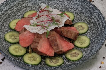 Exquisite medium-rare roast beef salad with cucumber and garnish in bowl, angled on background.