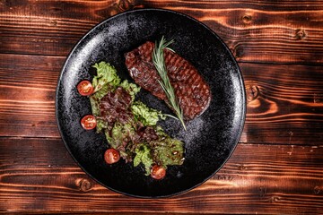 Appetizing Grilled Steak with Rosemary and Fresh Salad on Black Plate, Showcasing an Exquisite Blend of Flavors and Textures on a Wooden Table, Top View.