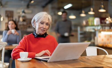 Senior woman cafe visitor sits at table with laptop, reads incoming emails and drinks strong...