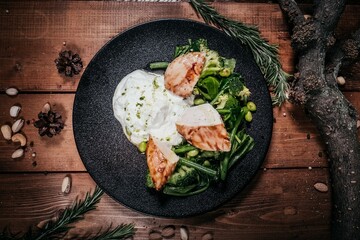 Premium Grilled Chicken with Creamy Sauce, Greens and Vegetables on Black Plate, Wood, Nuts and Herbs Decor, Serving on Wooden Table, Top View, Gastronomic Concept.