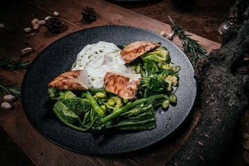 Signature Grilled Chicken with Cream and Green Vegetables on Black Plate, Served with Decoration of Wood, Nuts and Rosemary Branches on Wooden Table, Top View.