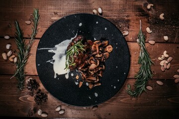 Top View: Roasted Lamb Shank with Forest Mushrooms on Black Plate, Decorated with Rosemary Sprigs, Wooden Table Decor, Culinary Splendor and Gourmet Treat.