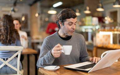 Man cafe visitor sits at table with laptop, reads incoming emails and drinks strong coffee. Client is preparing for important meeting, reading text content of report