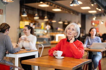 Cafe visitor drinks coffee alone. Senior woman whiles away time and waits for meeting in chain cafe, spend time while drinking coffee.