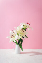White lilies in a ceramic pitcher placed against a pink background. Closeup