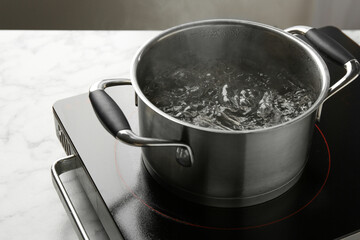Cooking pot with boiling water and stove on white marble table against grey background, closeup