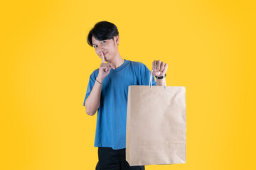 Portrait of young man showing paper bag and finger on lips asking for silence with playful...