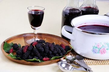 Deep red fruit juice in a long stem glass is placed next to mulberries on a wooden tray and is...