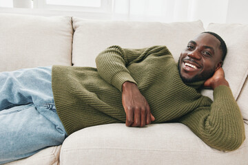 Obraz premium Smiling Black man relaxing on a cozy sofa in stylish green sweater, showing happiness and comfort in a bright, modern living room