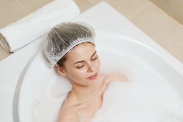 Relaxed young woman enjoying a bubble bath in a modern tub, wearing a shower cap, indulging in self-care and wellness at home. Serenity and relaxation concept.