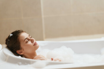 Naklejka premium Relaxed young woman enjoying a bubble bath, her eyes closed, embracing tranquility and self-care in a serene bathroom setting. Wellness and relaxation concept.