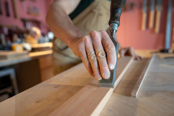 Experienced carpenter hands skillfully smoothing wood board surface with precise chisel removing imperfections. Detailed manual tool work, wood finishing sanding, surface refinement, craftsmanship