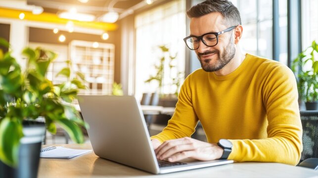 Bright cheerful man wea glasses and a yellow shirt working on a laptop in a modern cozy cafe with large and lush green plants for a relaxed work environment