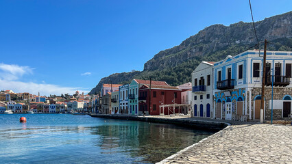 Obraz premium Colorful Waterfront Houses In Kastellorizo, Greece, Lined Along A Calm Harbor With Mountain Backdrop, Bright Sunny Day, Concept Of Mediterranean Travel, Real Estate, Summer Vacation, Tourism Promotion