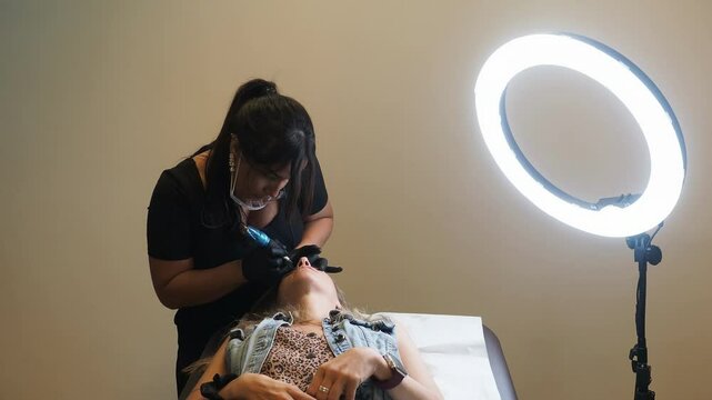 Client reclines on table while esthetician performs eyebrow microblading under bright ring light, hands in action, full setup visible including tool and studio elements &mdash; slow motion.