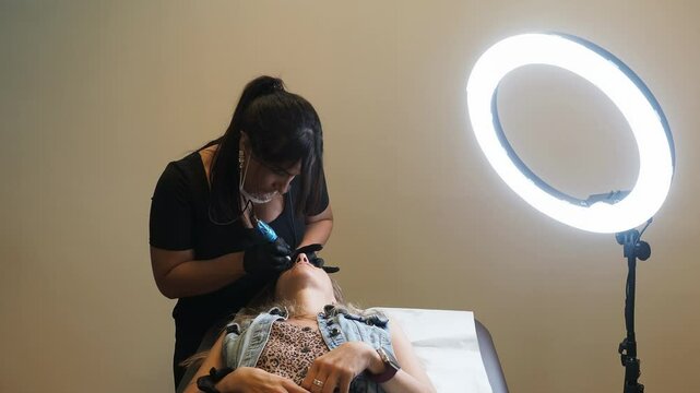 Esthetician bends over client lying on treatment chair while performing eyebrow microblading under ring light, both subjects framed fully with visible tool and studio setup &mdash; slow motion.