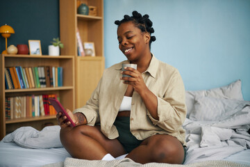 African American woman sitting on bed, holding smartphone and beverage, smiling in a cozy bedroom...