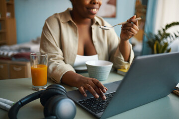 Smiling woman sitting at kitchen table with laptop, glass of juice, and headphones, enjoying breakfast and browsing on laptop in comfortable setting