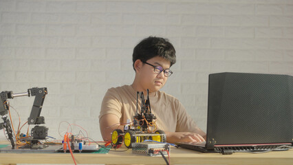 Asian teenage boy concentrating on coding a robotics project using a laptop, with STEM tools and circuits on the side, highlighting modern education in technology and engineering.
