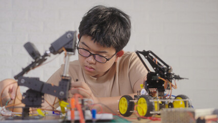 Asian teenage boy connecting wires to circuit board of robotic car with robotic arm at home, STEM education, robotics, and coding for young engineers.