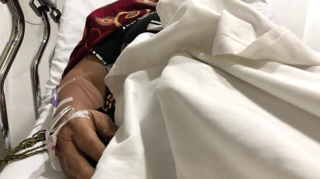 mature woman lies in a hospital bed with a dextrose IV attached to her right hand.