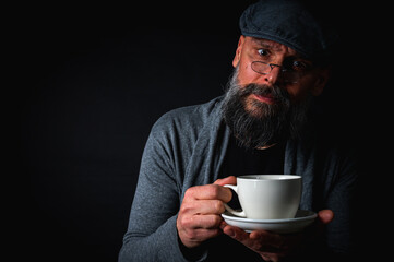 Stylish bearded man in cap and glasses holding coffee cup and saucer on dark background. Thoughtful expression, cozy casual wear, modern vintage atmosphere, morning tea drinking