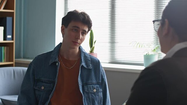 Slowmo medium shot of vulnerable Caucasian teen boy getting emotional while opening up about his problems to female psychologist sitting in front of him, counseling during therapy consultation