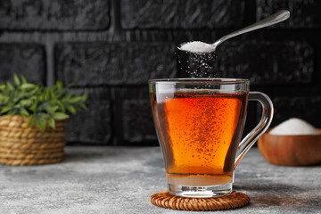 White sugar falling from teaspoon into glass cup of hot tea placed on coaster against black brick wall as background