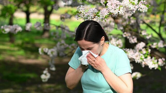 Young woman sneezing and wiping tears with tissue in blooming sakura garden, showing signs of seasonal allergy and hay fever