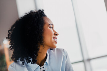 Obraz premium Young woman smiling brightly near a large window in a cozy living room on a sunny day, embracing relaxation and a positive lifestyle.