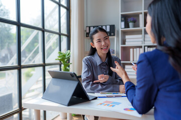 Two asian businesswomen are discussing work, using a tablet and charts in a modern office with large windows, collaborating on a project and sharing ideas