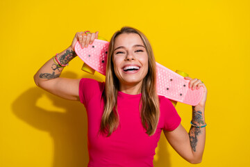 Smiling young woman holding pink skateboard on yellow backdrop showcasing trendy summer fashion and...