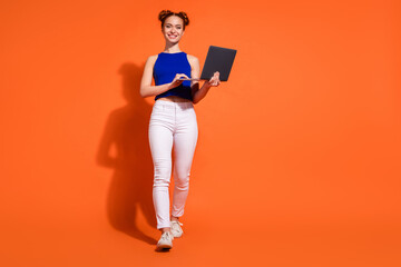 Youthful redheaded woman holding laptop, smiling on orange background, white pants and blue top, emphasizing casual fashion and technology