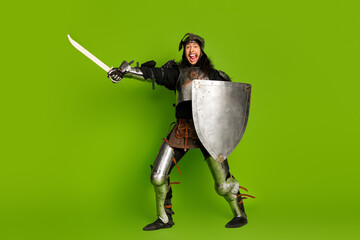 Energetic man in medieval armor with sword and shield posing on vibrant green background