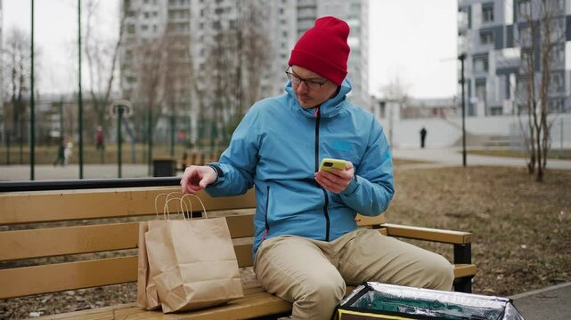 Food delivery man wearing casual clothes and red winter hat is packing food order into thermal bag while sitting on bench in city park, and using smartphone app