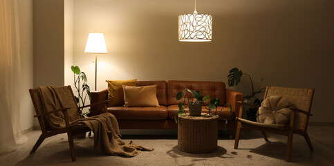 Interior of living room with brown sofa, armchairs, table and glowing lamps in evening