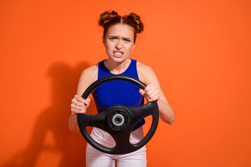 Portrait of a young woman with a funny expression holding a car steering wheel, posing against a...