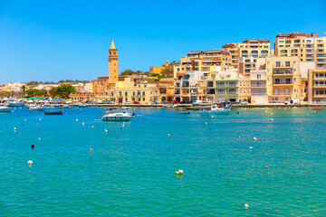Colorful waterfront view of Marsaskala, Malta. Boats gently float in turquoise waters against a...