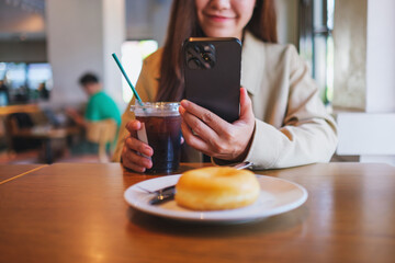 Closeup of a woman holding and using mobile phone while drinking iced coffee and eating donut at coffee shop
