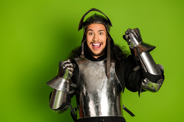 Excited young man in shining medieval knight armor poses against a vibrant green background
