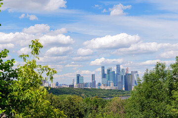 Fototapeta premium Breathtaking view of Moscow skyline from Vorobyovy Gory, with Moscow River, green trees, and clouds in the blue sky. The Moscow skyline dominates the horizon, creating a stunning scene
