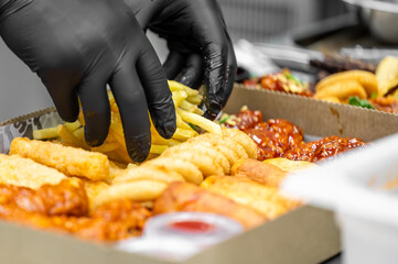 Hands in black gloves arrange fried snacks including French fries, onion rings, and chicken wings...