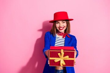 Smiling fashionable young woman in blue blazer and red hat presenting gift box on pink background, celebrating holiday season
