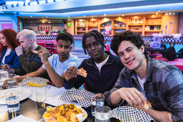 Group of diverse young friends enjoying pizza and nachos at a bowling alley restaurant, sharing...